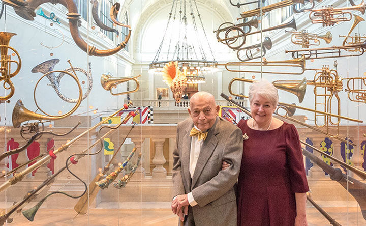 A man and woman smiling, standing in a gallery with horns and other brass instruments around them, flags and a chandelier are visible in the background.