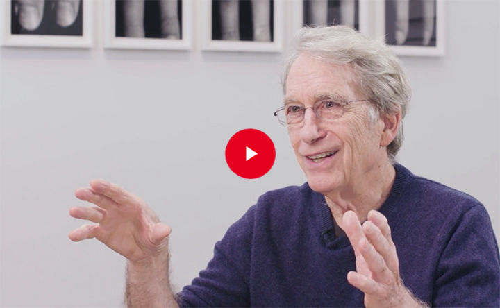 Still image from a video of a man wearing glasses, speaking in a gallery with photographs on the wall.