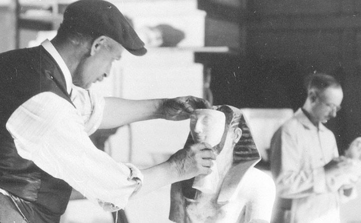 Detail of archival black-and-white photograph showing a man in a white shirt and black vest wearing a black cap fitting the face onto an Egyptian head