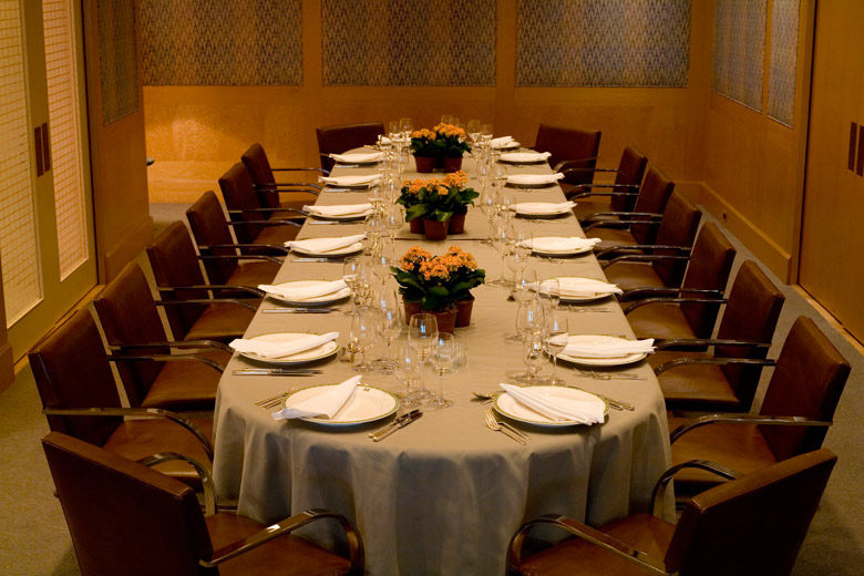 A casual-elegant private dining room with blond wood paneling and fabric covered walls, leather and chrome chairs; the table is set for eighteen with a casual service and low, elegant flower arrangements