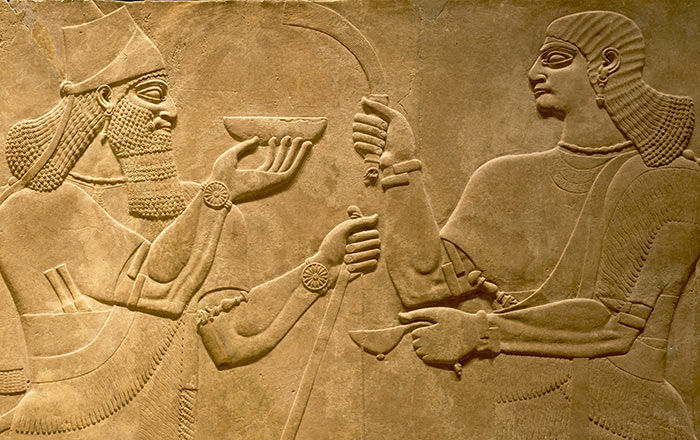 A close-up of a large stone relief panels depicting a long-haired bearded king in a conical cap with a small peak and a long diadem (the royal crown); he holds a bow, and a ceremonial bowl; facing him, is a beardless man carrying a fly whisk and a ladle